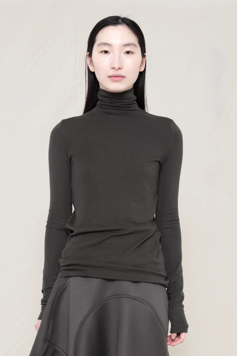 woman-wearing-a-warm-grey-soft-fluid-rib-long-sleeve-turtleneck-with-an-mb-charm-necklace-over-a-grey-spongy-seamed-skirt-and-custom-shoes-against-a-white-background-front-view