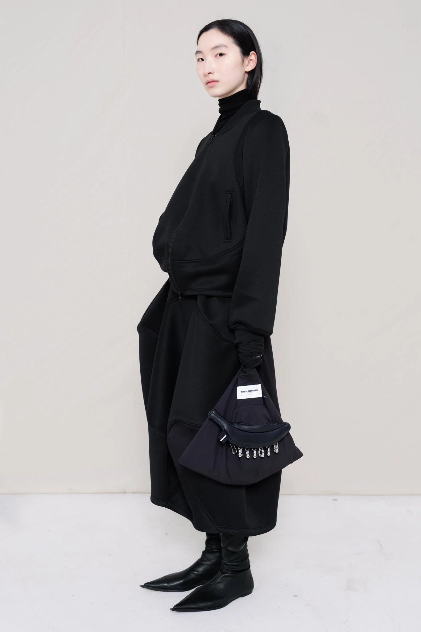 PERSON-WEARING-BLACK-WAVY-ZIP-UP-WITH-MATCHING-WAVY-SEAM-SKIRT-AND-BLACK-FLAT-POINTY-BOOTS-HOLDING-BLACK-PIERCED-BANANA-TOTE-BAG-THREE-QUARTER-VIEW
