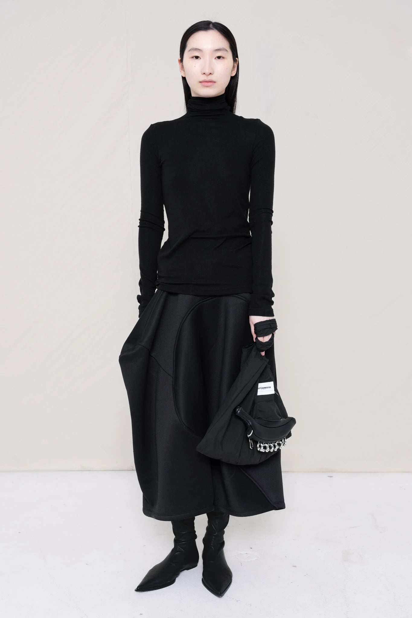 woman-wearing-a-black-soft-fluid-rib-long-sleeve-turtleneck-with-an-mb-charm-necklace-over-a-black-spongy-seamed-skirt-black-pointed-leather-boots-and-black-banana-tote-against-a-white-background-full-body-front-view