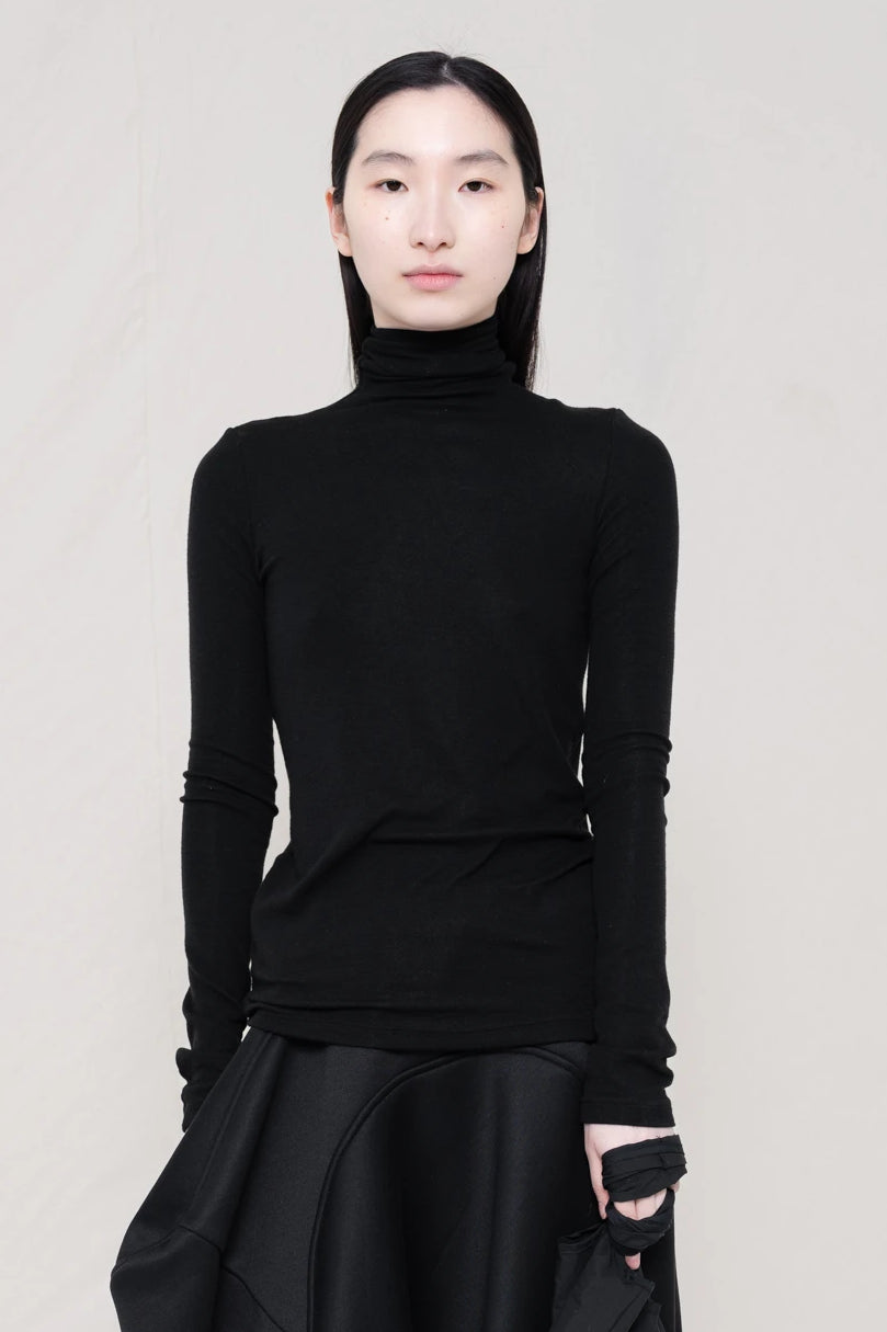 woman-wearing-a-black-soft-fluid-rib-long-sleeve-turtleneck-with-an-mb-charm-necklace-over-a-black-spongy-seamed-skirt-black-pointed-leather-boots-and-black-banana-tote-against-a-white-background-front-view