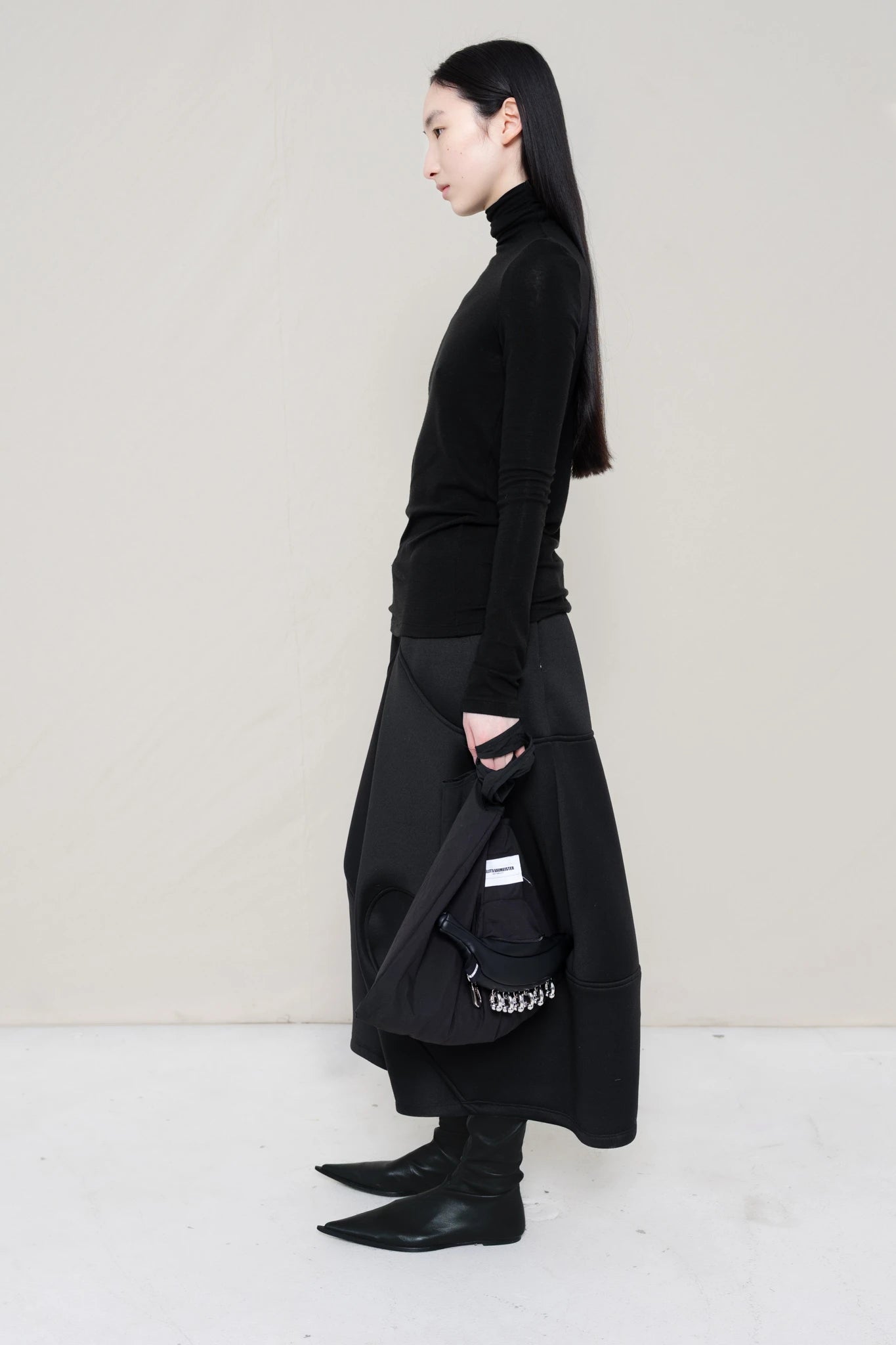 woman-wearing-a-black-soft-fluid-rib-long-sleeve-turtleneck-with-an-mb-charm-necklace-over-a-black-spongy-seamed-skirt-black-pointed-leather-boots-and-black-banana-tote-against-a-white-background-full-body-side-view