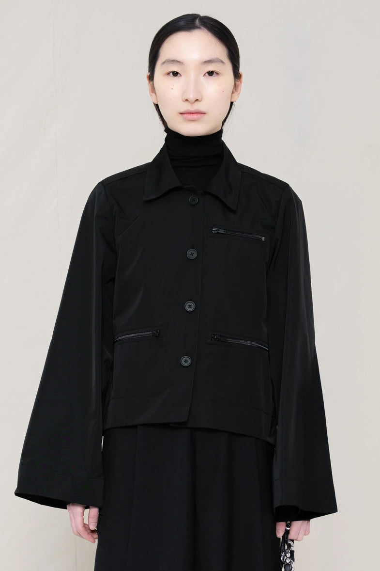 woman-wearing-boxy-jacket-fit-black-taffeta-shirt-with-collar-zipper-pockets-and-wide-sleeves-over-black-turtle-neck-and-black-wide-trousers-against-a-plain-background-front-view