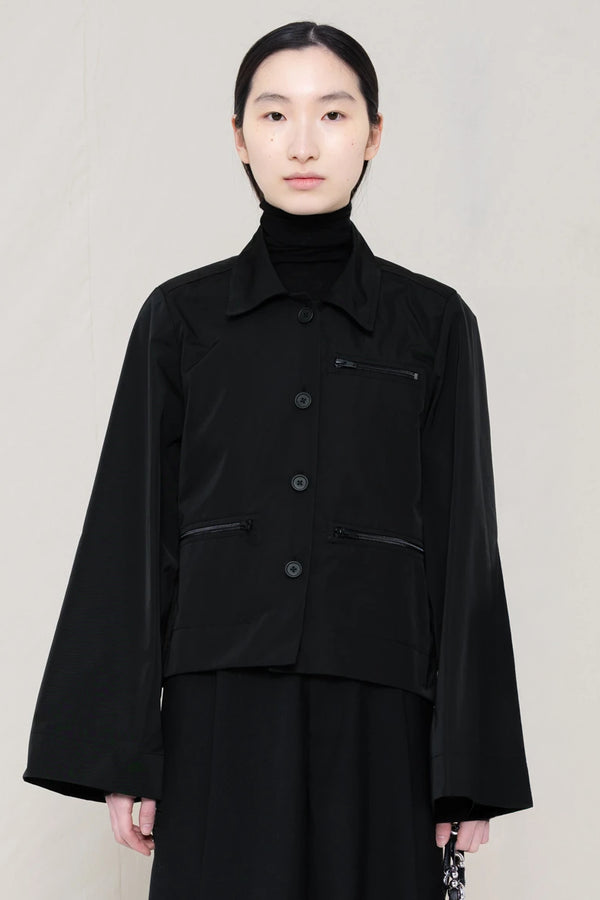 woman-wearing-boxy-jacket-fit-black-taffeta-shirt-with-collar-zipper-pockets-and-wide-sleeves-over-black-turtle-neck-and-black-wide-trousers-against-a-plain-background-front-view
