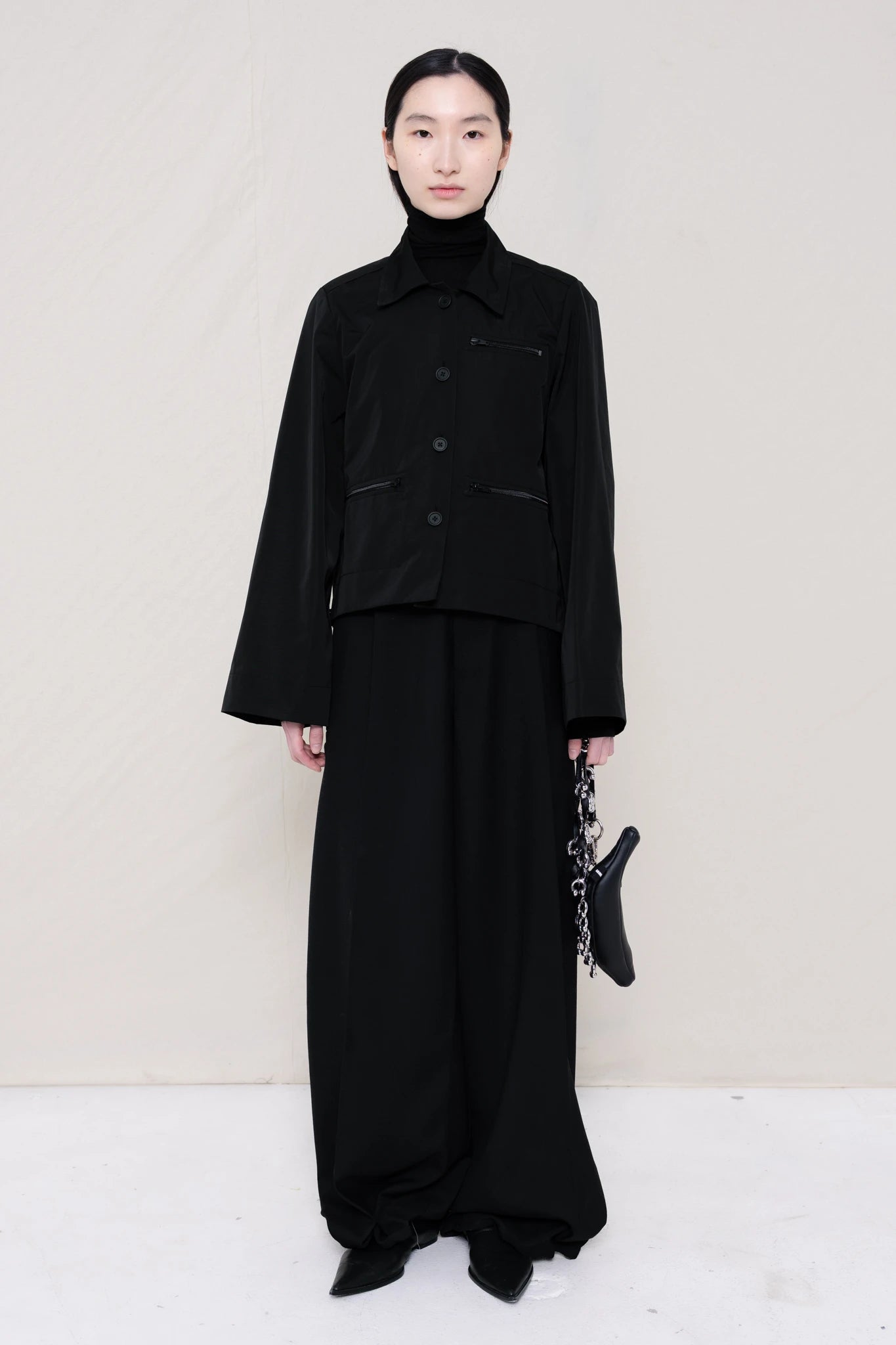 woman-holding-a-black-leather-banana-bag-with-a-studded-strap-wearing-boxy-jacket-fit-black-taffeta-shirt-with-collar-zipper-pockets-and-wide-sleeves-over-black-turtle-neck-and-black-wide-trousers-with-black-leather-pointed-boots-against-a-plain-background-full-body-front-view