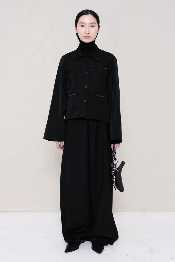 woman-holding-a-black-leather-banana-bag-with-a-studded-strap-wearing-boxy-jacket-fit-black-taffeta-shirt-with-collar-zipper-pockets-and-wide-sleeves-over-black-turtle-neck-and-black-wide-trousers-with-black-leather-pointed-boots-against-a-plain-background-full-body-front-view