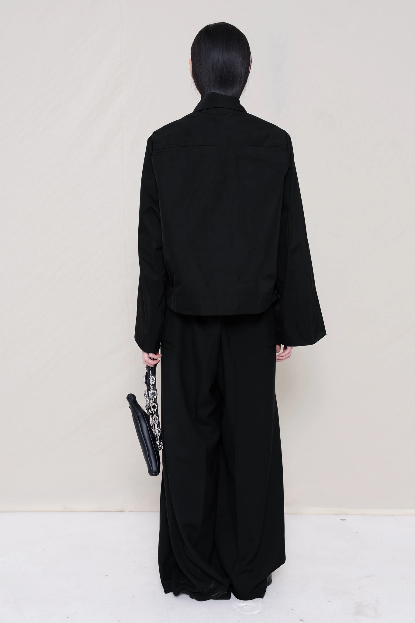 woman-holding-a-black-leather-banana-bag-with-a-studded-strap-wearing-boxy-jacket-fit-black-taffeta-shirt-with-collar-zipper-pockets-and-wide-sleeves-over-black-turtle-neck-and-black-wide-trousers-with-black-leather-pointed-boots-against-a-plain-background-full-body-back-view