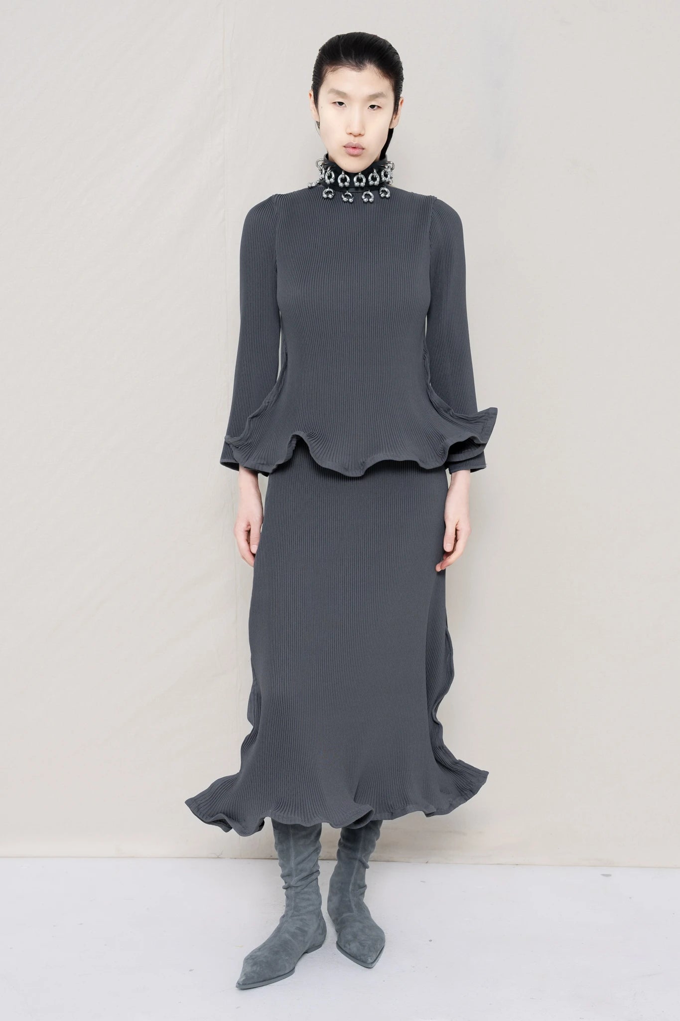 PERSON-WEARING-COLD-GREY-RIPPLE-PLEAT-ZIG-ZAG-HEM-TOP-AND-MATCHING-RIPPLE-PLEAT-ZIG-ZAG-HEM-SKIRT-ACCESSORIZED-WITH-PIERCED-CHOKER-AND-GREY-FLAT-POINTY-BOOTS-FRONT-VIEW
