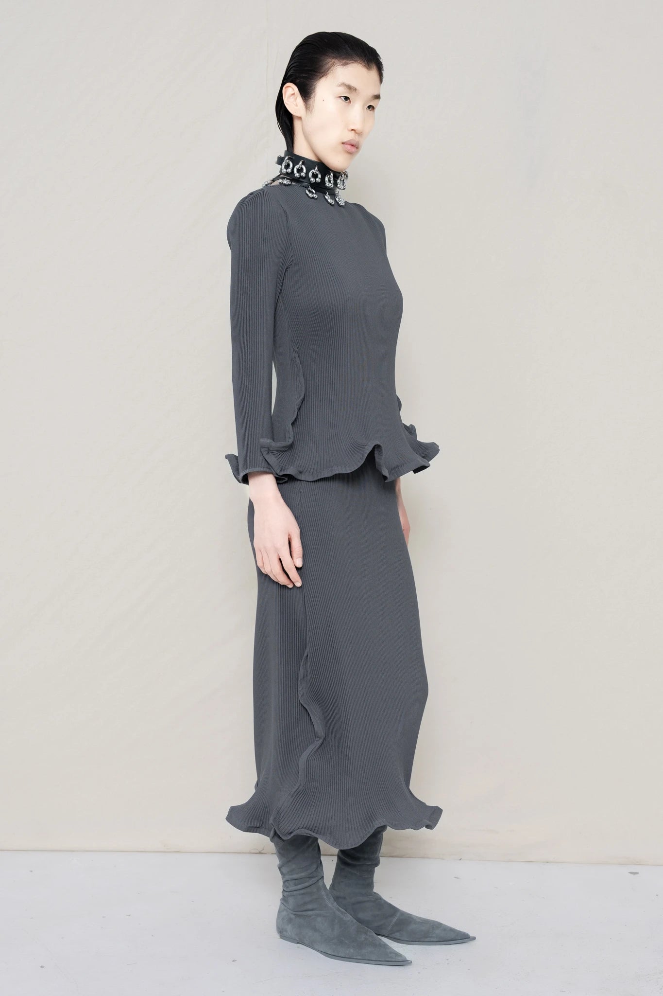 PERSON-WEARING-COLD-GREY-RIPPLE-PLEAT-ZIG-ZAG-HEM-TOP-AND-MATCHING-RIPPLE-PLEAT-ZIG-ZAG-HEM-SKIRT-ACCESSORIZED-WITH-PIERCED-CHOKER-AND-GREY-FLAT-POINTY-BOOTS-THREE-QUARTER-VIEW