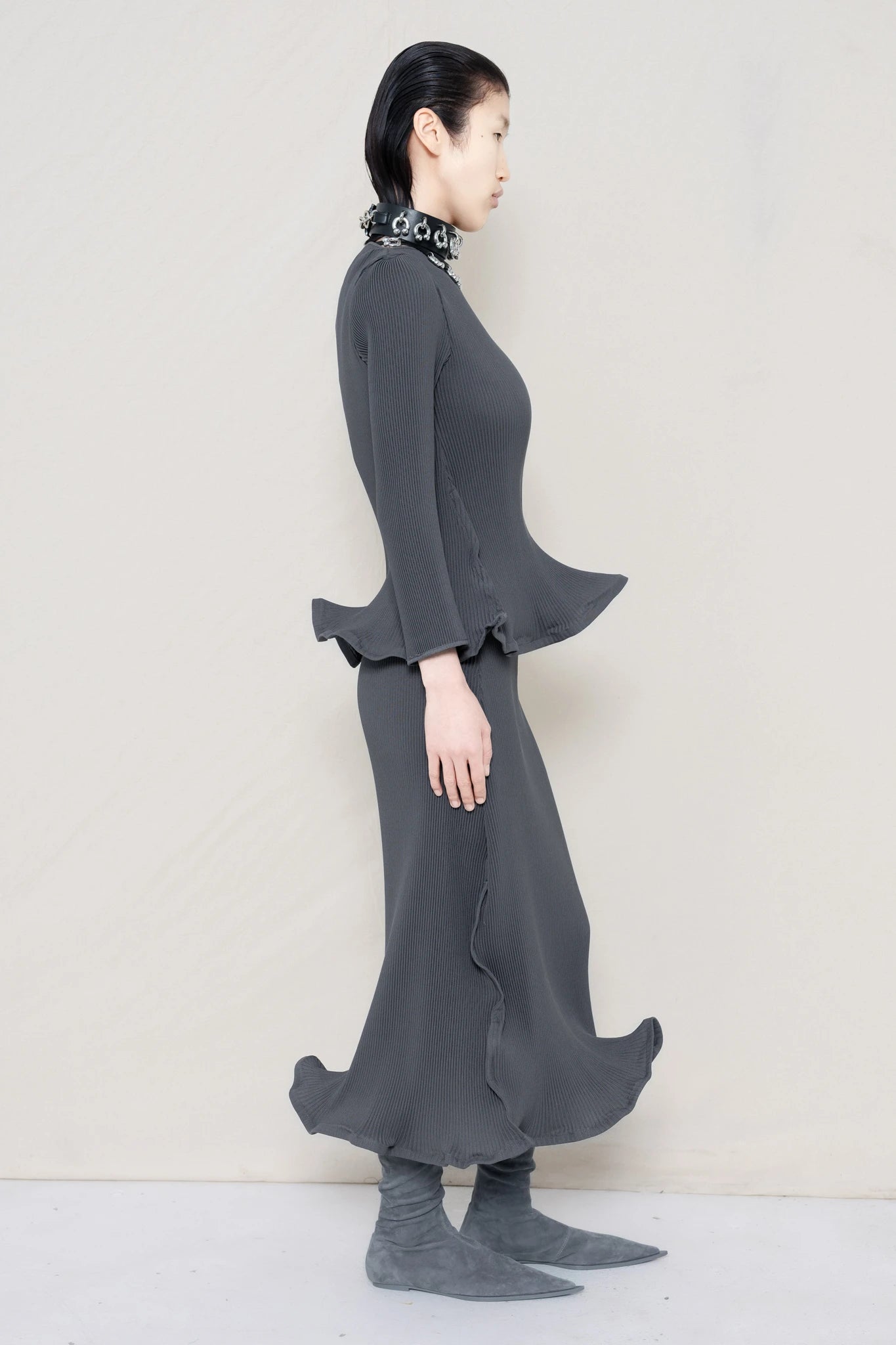 PERSON-WEARING-COLD-GREY-RIPPLE-PLEAT-ZIG-ZAG-HEM-TOP-AND-MATCHING-RIPPLE-PLEAT-ZIG-ZAG-HEM-SKIRT-ACCESSORIZED-WITH-PIERCED-CHOKER-AND-GREY-FLAT-POINTY-BOOTS-SIDE-VIEW