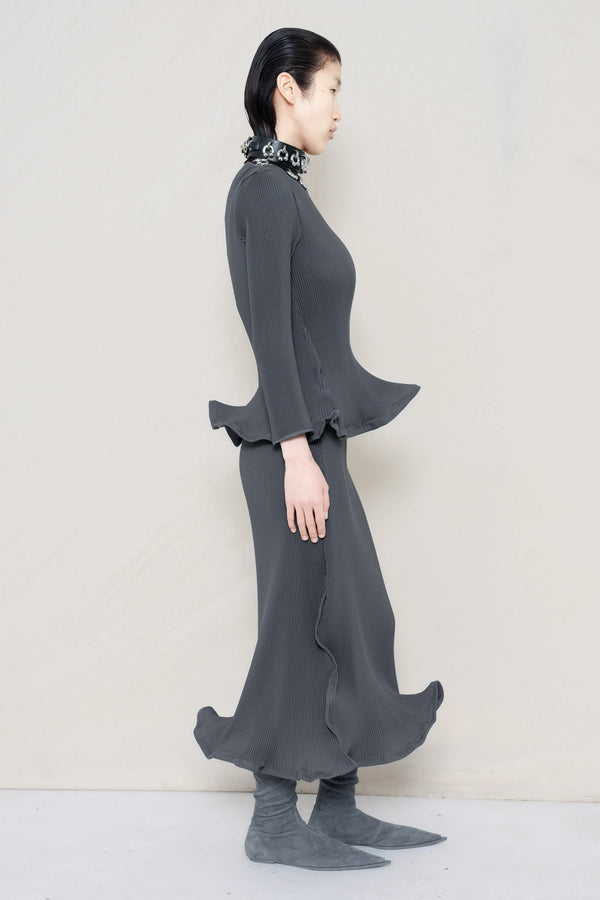 PERSON-WEARING-COLD-GREY-RIPPLE-PLEAT-ZIG-ZAG-HEM-TOP-AND-MATCHING-RIPPLE-PLEAT-ZIG-ZAG-HEM-SKIRT-ACCESSORIZED-WITH-PIERCED-CHOKER-AND-GREY-FLAT-POINTY-BOOTS-SIDE-VIEW