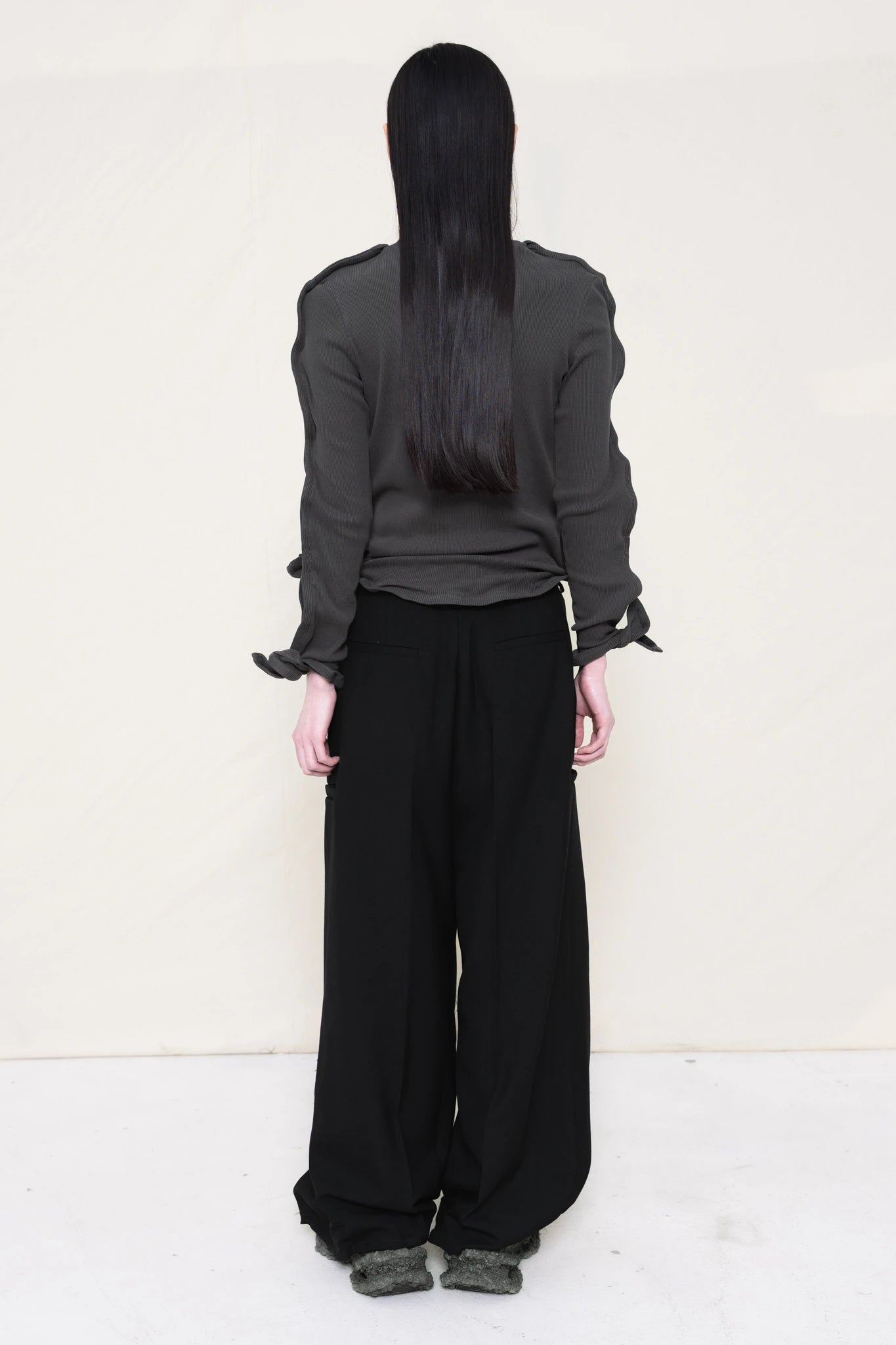 PERSON-WEARING-DARK-GREY-ZIG-ZAG-TOP-FEATURING-MALLEABLE-SLEEVES-PAIRED-WITH-BLACK-TAILORED-PANTS-AND-CUSTOMIZED-SNEAKERS-BACK-VIEW