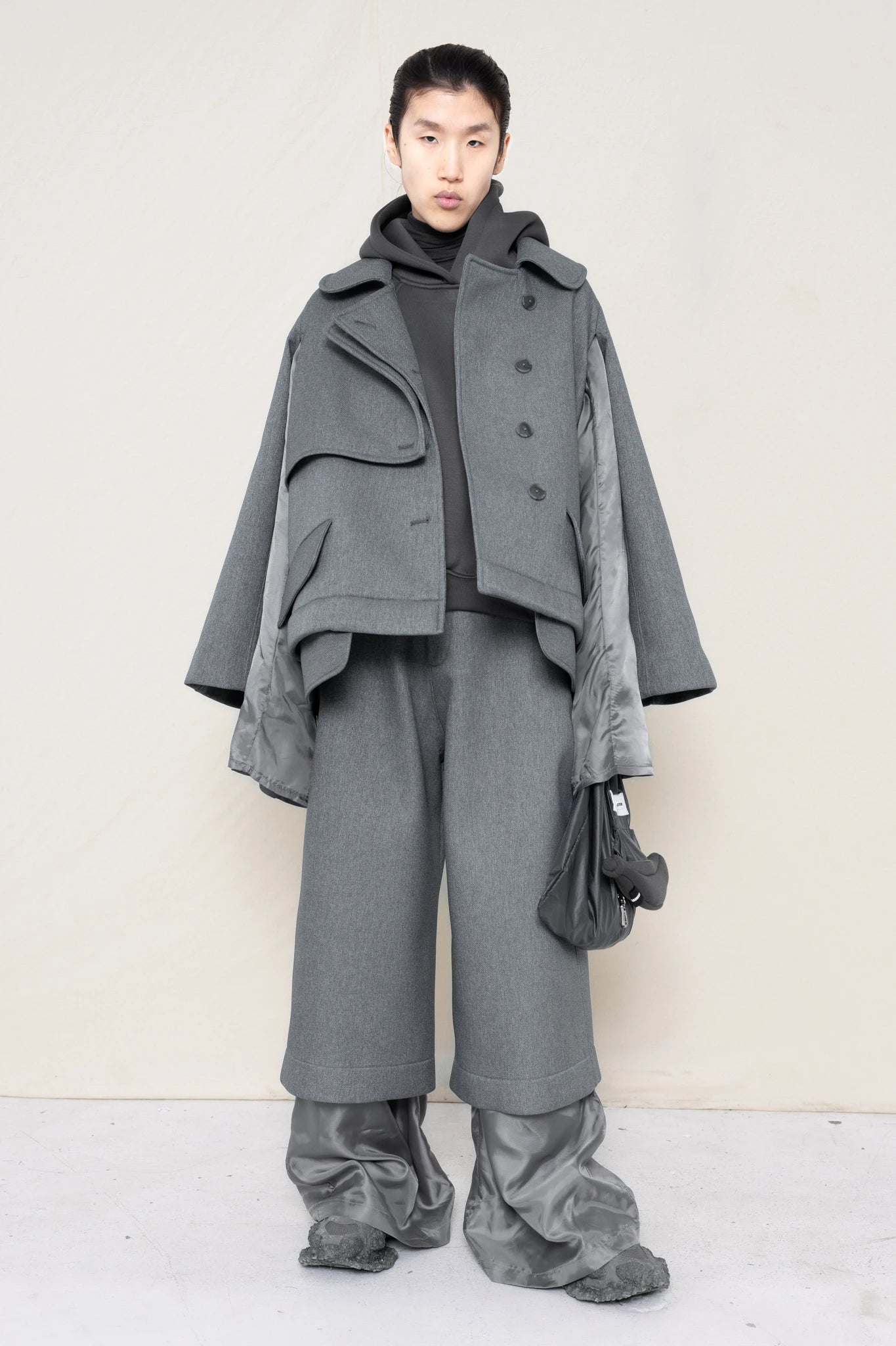 A-model-wearing-A-grey-DOUBLE-LAPEL-JACKET-WITH-grey-SILKY-SECOND-SLEEVEs-over-a-spongey-grey-hoodie-and-grey-trousers-and-silky-underlayer-draped-from-under-the-cuffs-AND-CUSTOMIZED-SNEAKERS-full-body-front-view