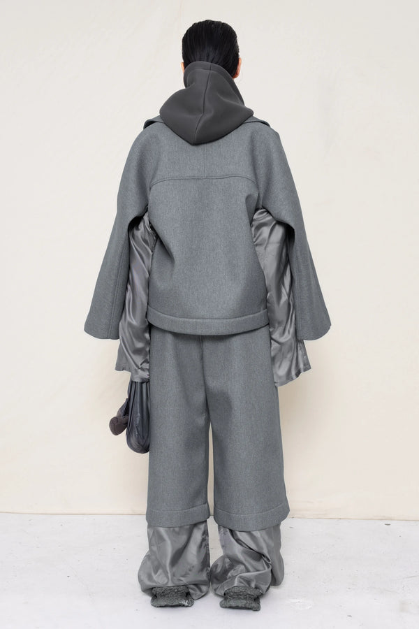 A-model-wearing-A-grey-DOUBLE-LAPEL-JACKET-WITH-grey-SILKY-SECOND-SLEEVEs-over-a-spongey-grey-hoodie-and-grey-trousers-and-silky-underlayer-draped-from-under-the-cuffs-AND-CUSTOMIZED-SNEAKERS-full-body-back-view