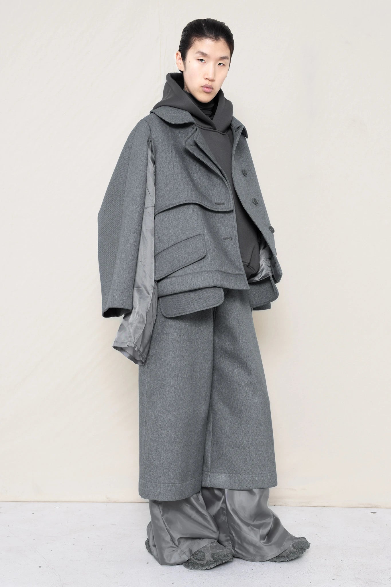 A-model-wearing-A-grey-DOUBLE-LAPEL-JACKET-WITH-grey-SILKY-SECOND-SLEEVEs-over-a-spongey-grey-hoodie-and-grey-trousers-and-silky-underlayer-draped-from-under-the-cuffs-AND-CUSTOMIZED-SNEAKERS-full-body-three-quarters-view