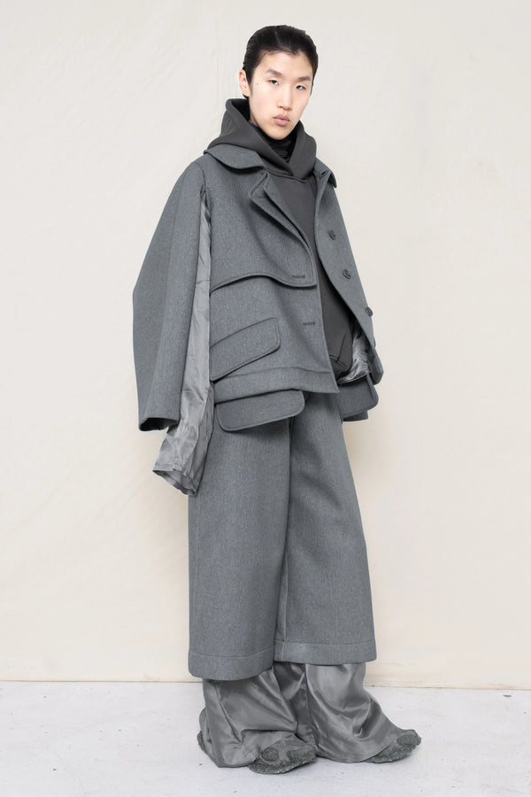 A-model-wearing-A-grey-DOUBLE-LAPEL-JACKET-WITH-grey-SILKY-SECOND-SLEEVEs-over-a-spongey-grey-hoodie-and-grey-trousers-and-silky-underlayer-draped-from-under-the-cuffs-AND-CUSTOMIZED-SNEAKERS-full-body-three-quarters-view