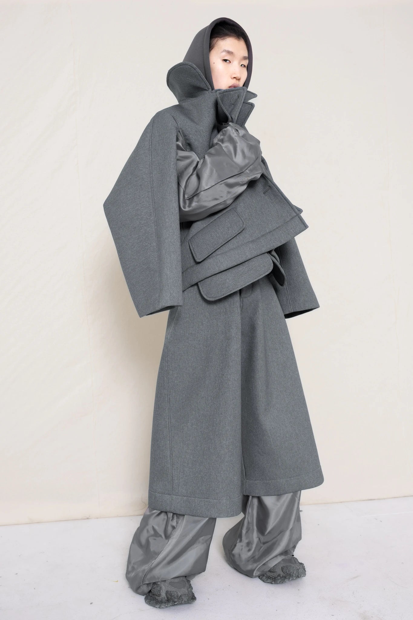 A-model-wearing-A-grey-DOUBLE-LAPEL-JACKET-WITH-grey-SILKY-SECOND-SLEEVEs-over-a-spongey-grey-hoodie-and-grey-trousers-and-silky-underlayer-draped-from-under-the-cuffs-full-body-three-quarters-view