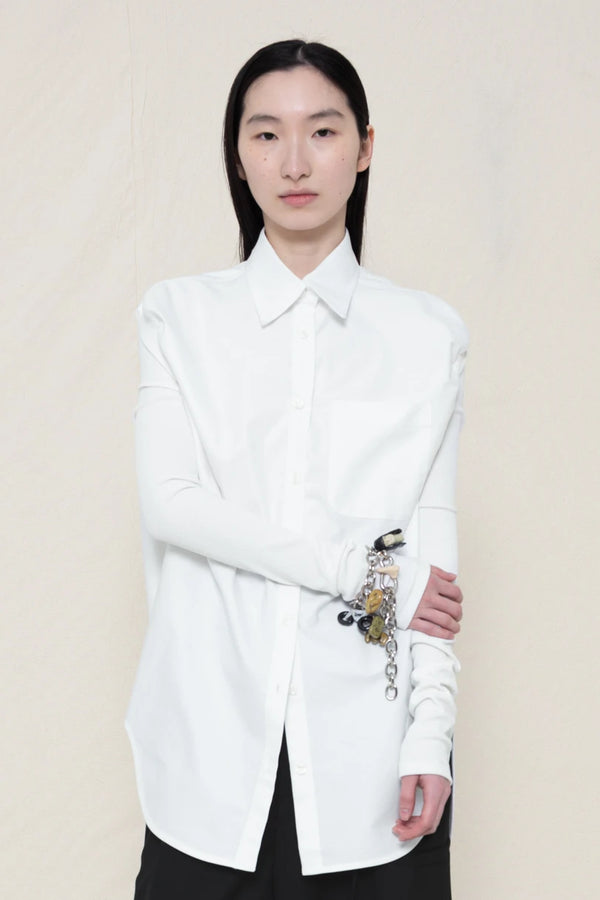 woman-wearing-a-slim-white-collared-button-down-shirt-with-ruched-shoulder-detail-over-wide-black-trousers-and-custom-shoes-and-a-charm-bracelet-against-a-plain-background-front-view