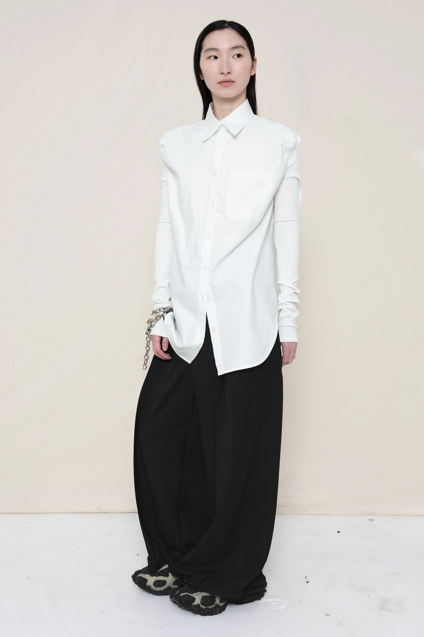 woman-wearing-a-slim-white-collared-button-down-shirt-with-ruched-shoulder-detail-over-wide-black-trousers-and-custom-shoes-and-a-charm-bracelet-against-a-plain-background-full-body-front-view