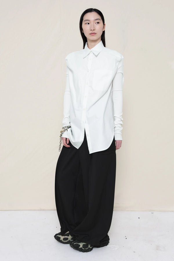 woman-wearing-a-slim-white-collared-button-down-shirt-with-ruched-shoulder-detail-over-wide-black-trousers-and-custom-shoes-and-a-charm-bracelet-against-a-plain-background-full-body-front-view