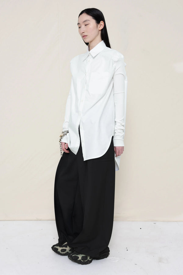 woman-wearing-a-slim-white-collared-button-down-shirt-with-ruched-shoulder-detail-over-wide-black-trousers-and-custom-shoes-and-a-charm-bracelet-against-a-plain-background-full-body-three-quarter-view