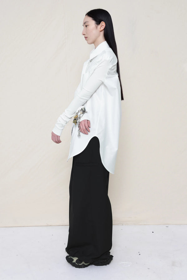 woman-wearing-a-slim-white-collared-button-down-shirt-with-ruched-shoulder-detail-over-wide-black-trousers-and-custom-shoes-and-a-charm-bracelet-against-a-plain-background-full-body-side-view