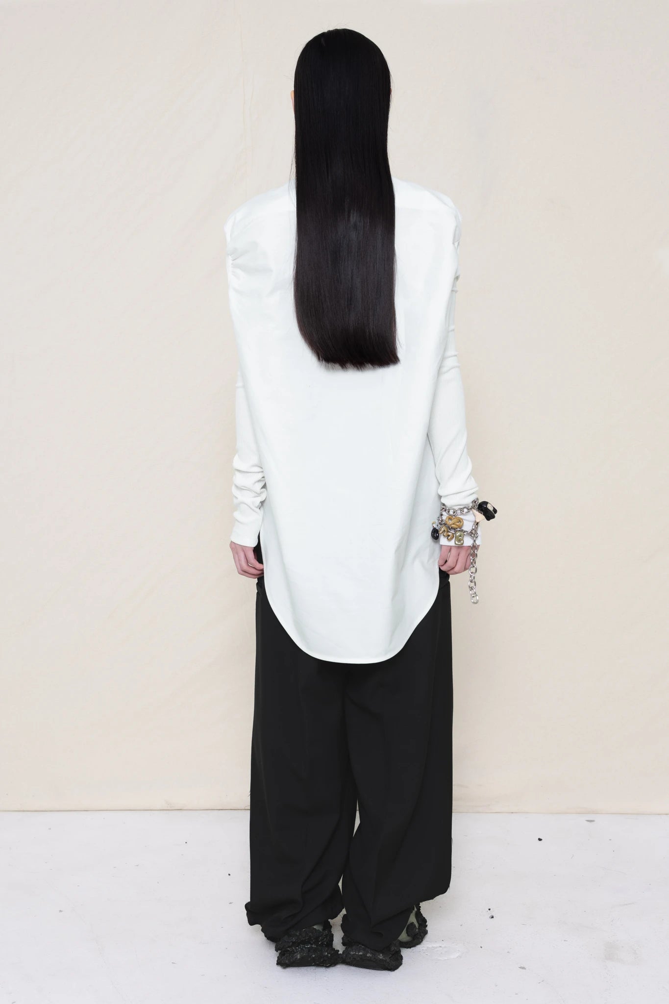 woman-wearing-a-slim-white-collared-button-down-shirt-with-ruched-shoulder-detail-over-wide-black-trousers-and-custom-shoes-and-a-charm-bracelet-against-a-plain-background-full-body-back-view