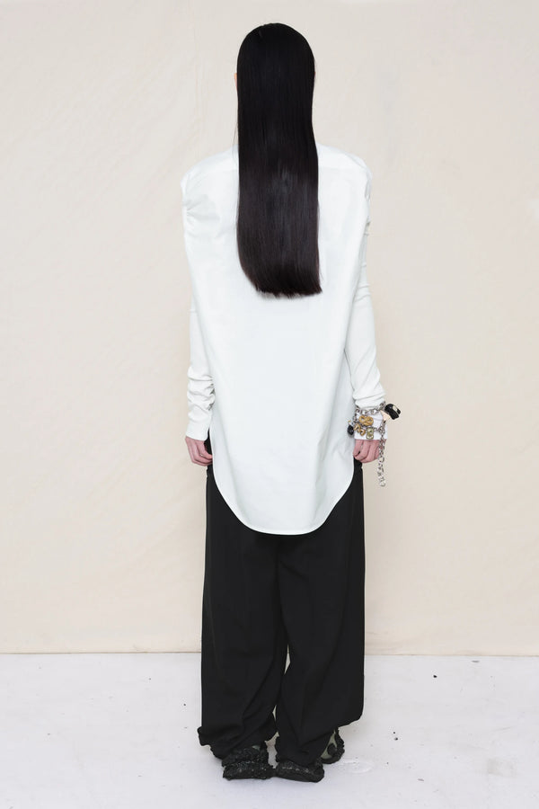 woman-wearing-a-slim-white-collared-button-down-shirt-with-ruched-shoulder-detail-over-wide-black-trousers-and-custom-shoes-and-a-charm-bracelet-against-a-plain-background-full-body-back-view