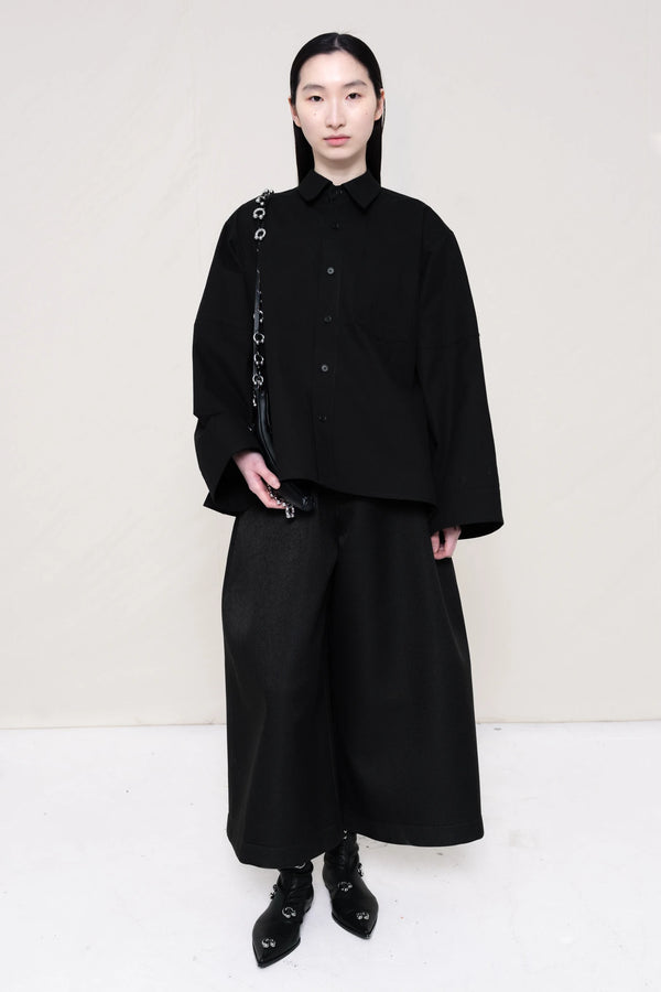 woman-wearing-a-black-boxy-fit-shirt-with-chest-pocket-and-wide-sleeve-cuffs-over-wide-black-trousers-and-black-leather-studded-pointed-boots-holding-a-black-leather-banana-bag-with-studded-strap-against-a-plain-background-full-body-front-view