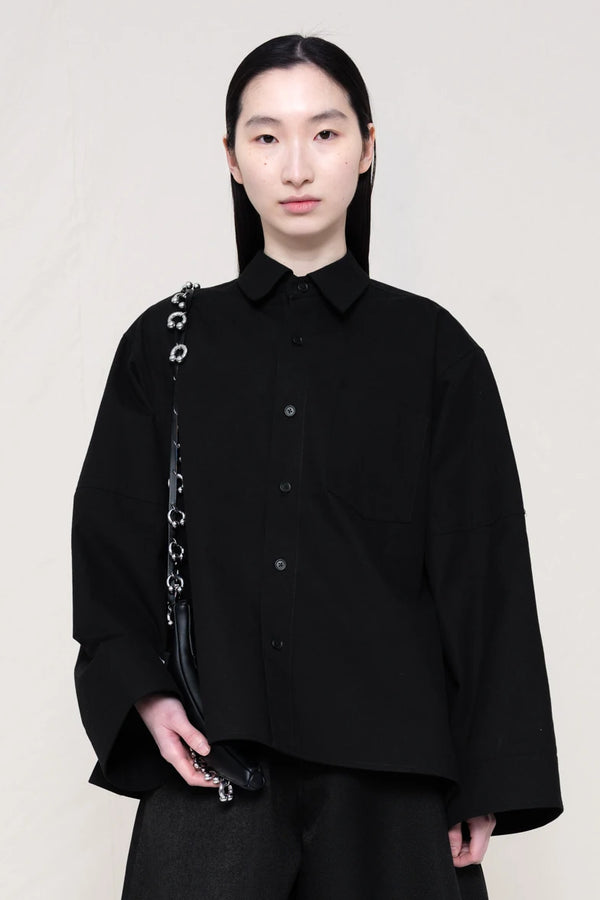 woman-wearing-a-black-boxy-fit-shirt-with-chest-pocket-and-wide-sleeve-cuffs-over-wide-black-trousers-holding-a-black-leather-banana-bag-with-studded-strap-against-a-plain-background-front-view