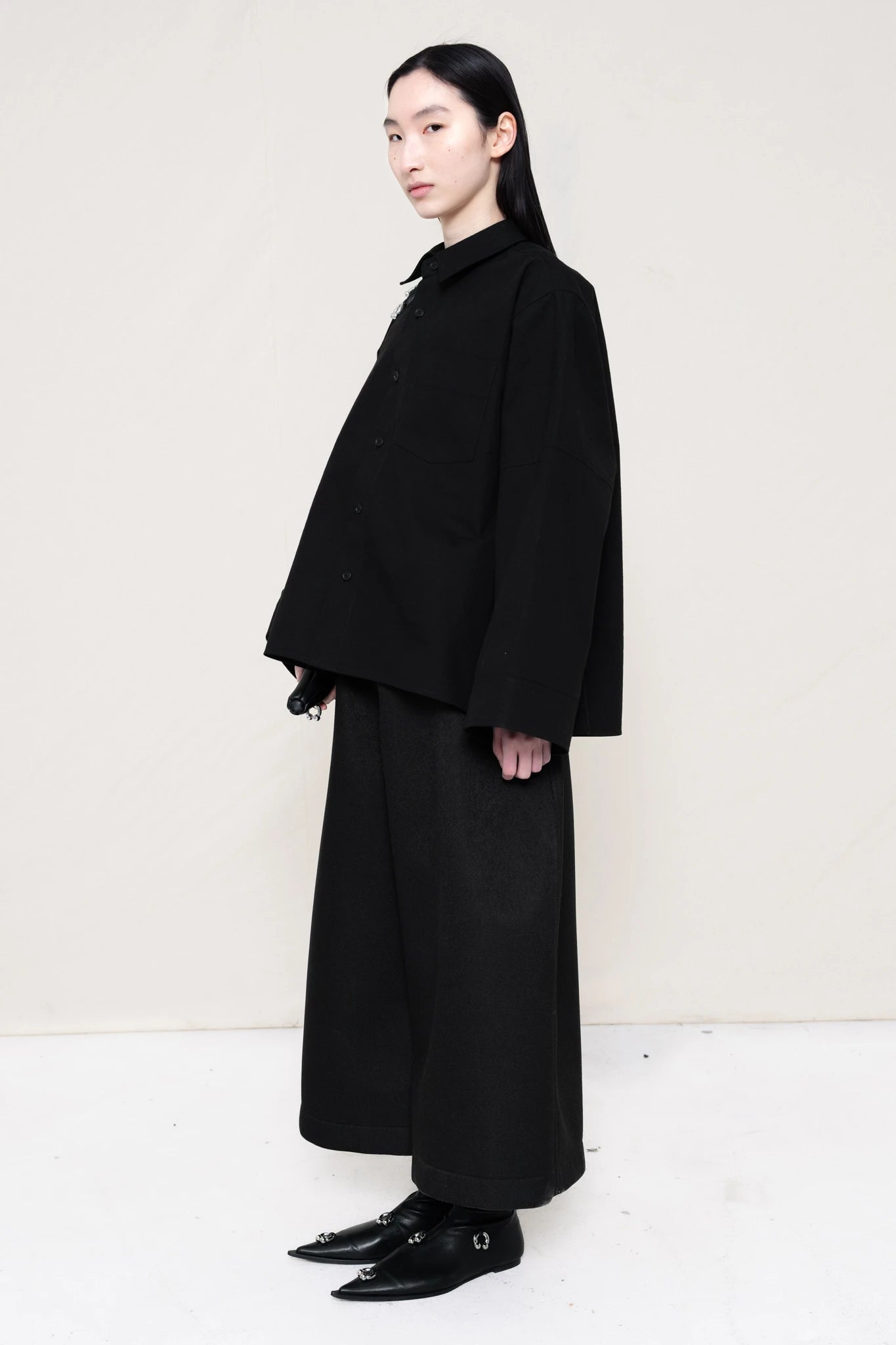 woman-wearing-a-black-boxy-fit-shirt-with-chest-pocket-and-wide-sleeve-cuffs-over-wide-black-trousers-and-black-leather-studded-pointed-boots-holding-a-black-leather-banana-bag-with-studded-strap-against-a-plain-background-full-body-three-quarters-view