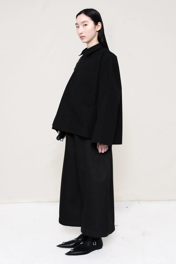 woman-wearing-a-black-boxy-fit-shirt-with-chest-pocket-and-wide-sleeve-cuffs-over-wide-black-trousers-and-black-leather-studded-pointed-boots-holding-a-black-leather-banana-bag-with-studded-strap-against-a-plain-background-full-body-three-quarters-view