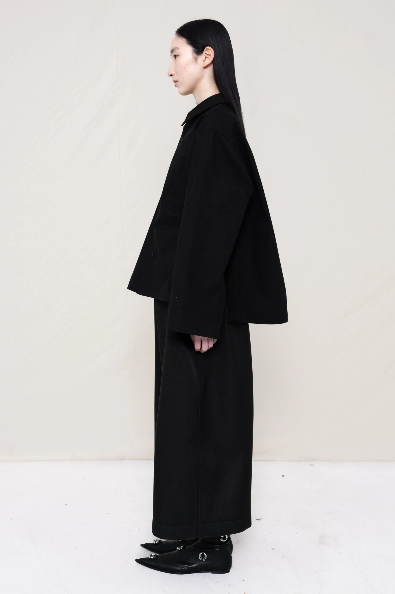 woman-wearing-a-black-boxy-fit-shirt-with-chest-pocket-and-wide-sleeve-cuffs-over-wide-black-trousers-and-black-leather-studded-pointed-boots-holding-a-black-leather-banana-bag-with-studded-strap-against-a-plain-background-full-body-side-view