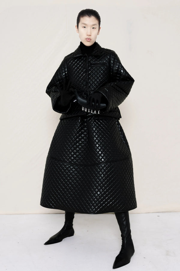 woman-wearing-a-black-quilted-jacket-over-a-black-quilted-a-line-calf-length-skirt-and-black-leather-pointed-boots-and-studded-black-leather-banana-bag-on-a-white-background-full-body-front-view