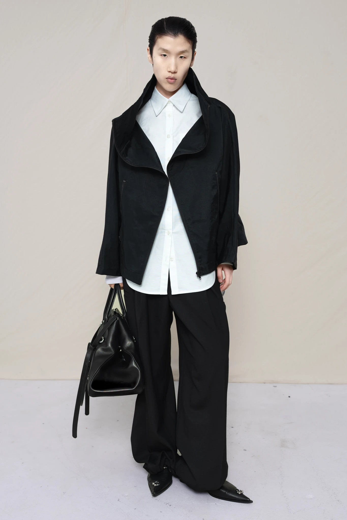 PERSON-WEARING-BLACK-BOXY-ZIP-UP-JACKET-WITH-WHITE-BUTTON-DOWN-AND-BLACK-TAILORED-PANTS-HOLDING-BLACK-PIERCED-A-LINE-BAG-WITH-MATCHING-PIERCED-FLAT-BOOTS-FRONT-VIEW
