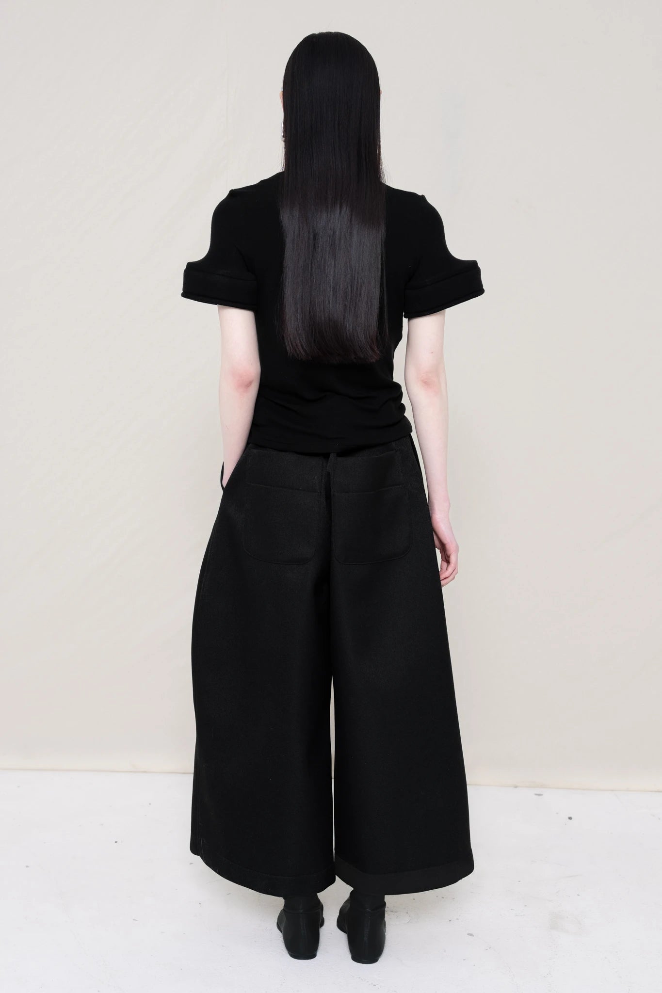 PERSON-WEARING-BLACK-TEE-WITH-FOAM-SLEEVE-CUFFS-AND-BLACK-CULOTTE-PANTS-WITH-BLACK-FLAT-POINTY-BOOTS-BACK-VIEW