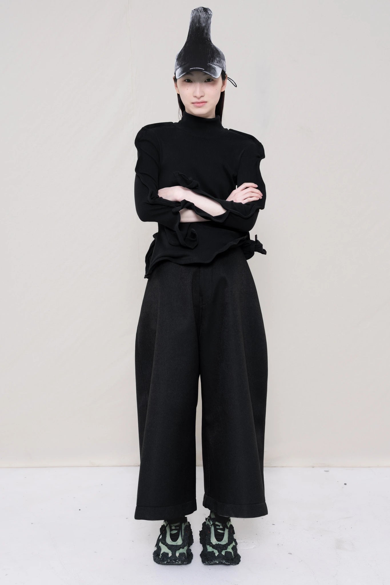PERSON-WEARING-PAINTED-PLEAT-POINTY-CAP-WITH-BLACK-ZIG-ZAG-TOP-FEATURING-MALLEABLE-SLEEVES-PAIRED-WITH-BLACK-CULOTTE-PANTS-AND-CUSTOMIZED-SNEAKERS-FRONT-VIEW