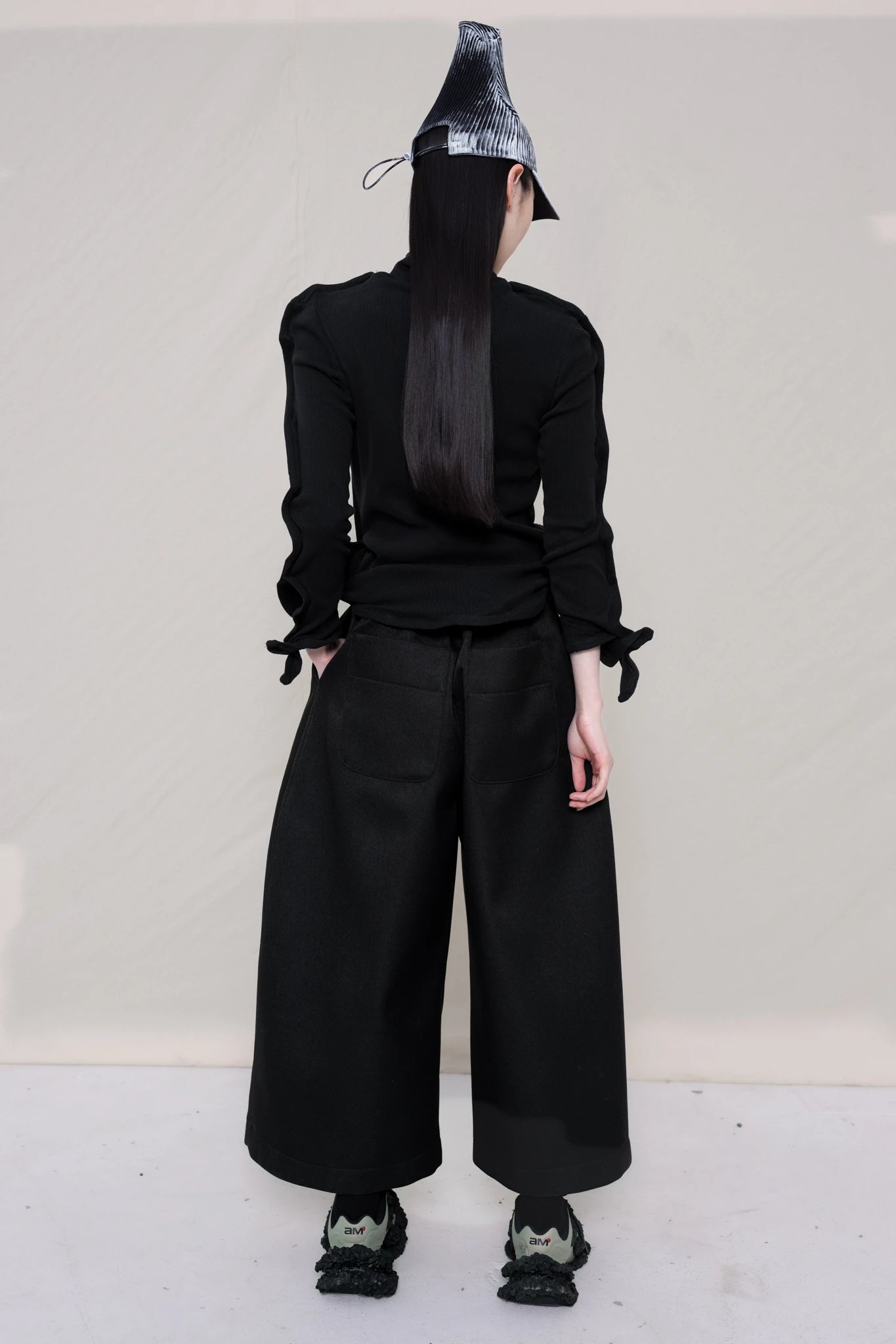 PERSON-WEARING-PAINTED-PLEAT-POINTY-CAP-WITH-BLACK-ZIG-ZAG-TOP-FEATURING-MALLEABLE-SLEEVES-PAIRED-WITH-BLACK-CULOTTE-PANTS-AND-CUSTOMIZED-SNEAKERS-BACK-VIEW