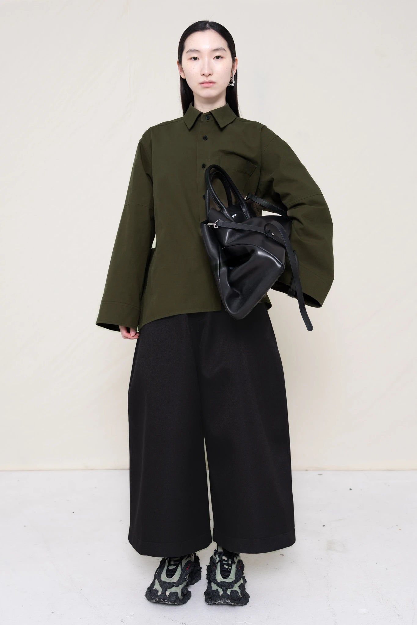 woman-wearing-a-moss-green-boxy-fit-shirt-with-chest-pocket-and-wide-sleeve-cuffs-over-wide-black-trousers-and-custom-shoes-holding-a-large-black-leather-bag-against-a-plain-background-full-body-front-view