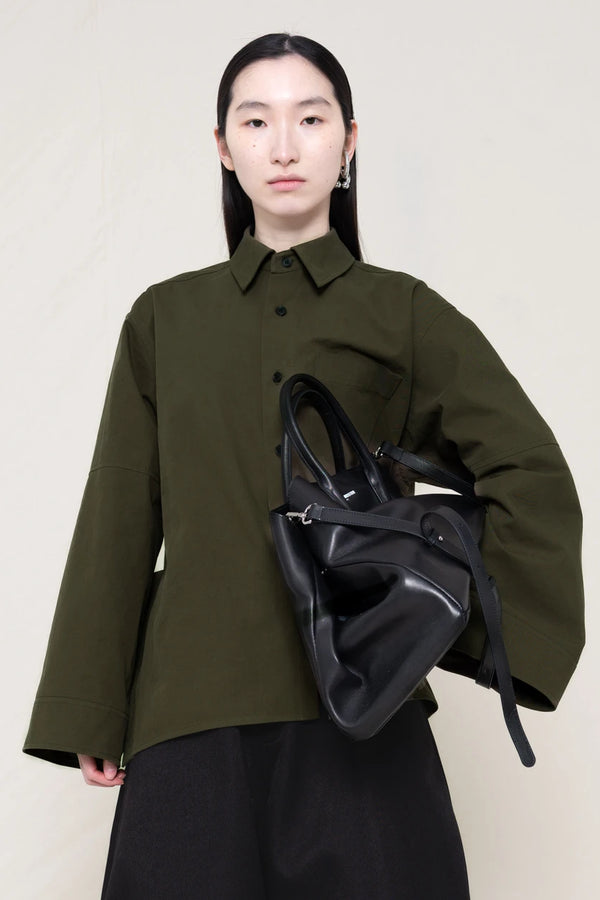 woman-wearing-a-moss-green-boxy-fit-shirt-with-chest-pocket-and-wide-sleeve-cuffs-over-wide-black-trousers-holding-a-large-black-leather-bag-against-a-plain-background-front-view
