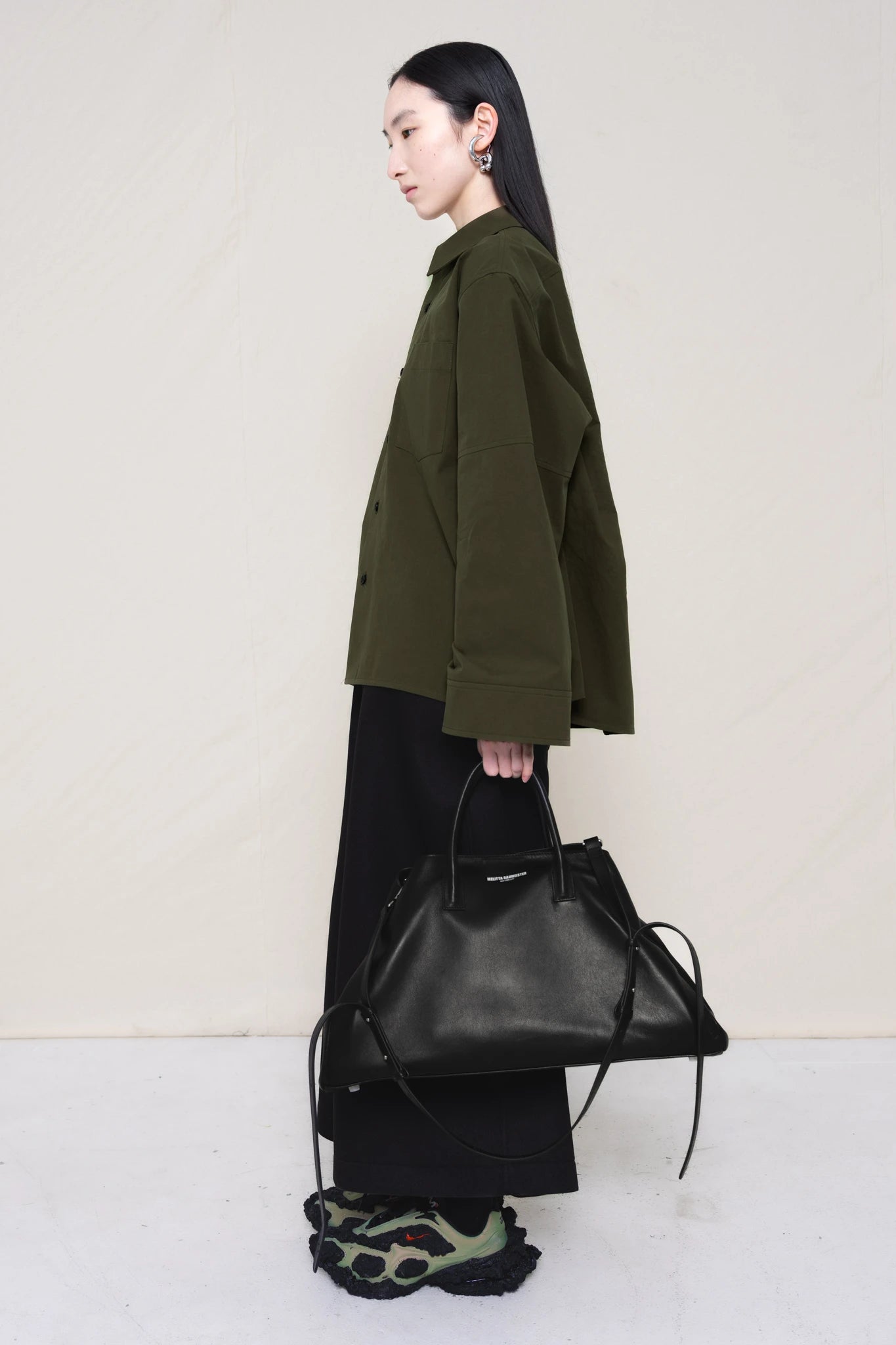 woman-wearing-a-moss-green-boxy-fit-shirt-with-chest-pocket-and-wide-sleeve-cuffs-over-wide-black-trousers-and-custom-shoes-holding-a-large-black-leather-bag-against-a-plain-background-full-body-side-view