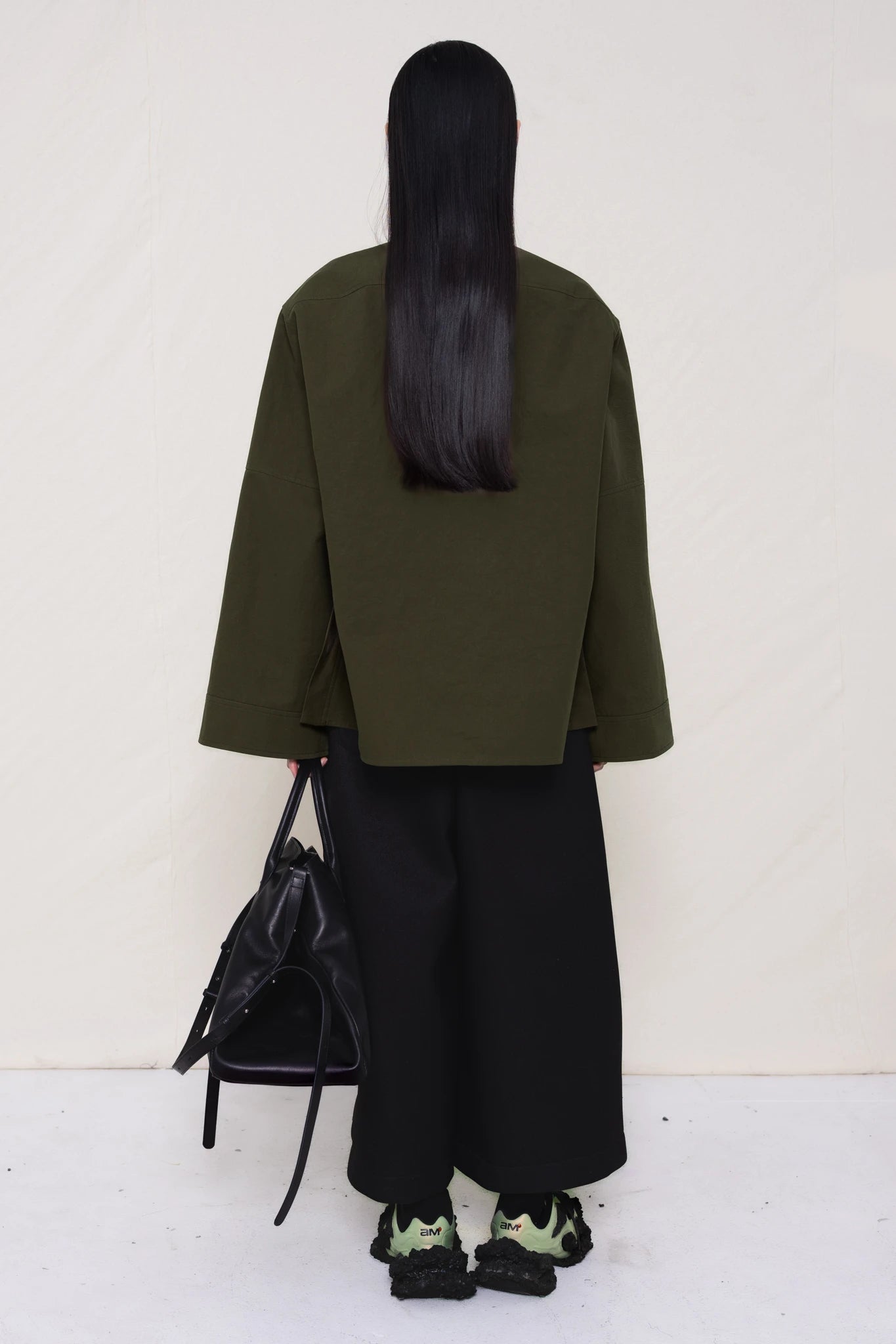 woman-wearing-a-moss-green-boxy-fit-shirt-with-chest-pocket-and-wide-sleeve-cuffs-over-wide-black-trousers-and-custom-shoes-holding-a-large-black-leather-bag-against-a-plain-background-full-body-back-view