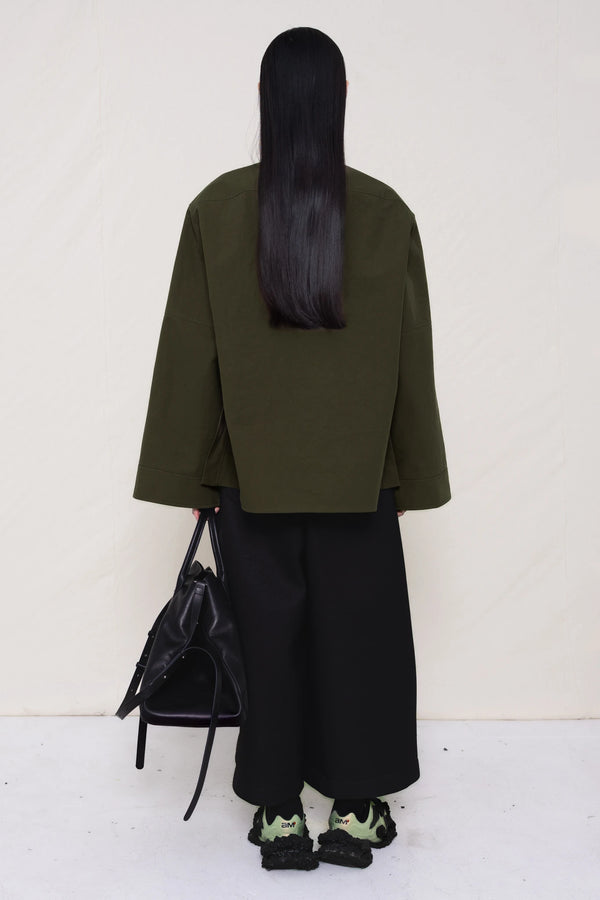 woman-wearing-a-moss-green-boxy-fit-shirt-with-chest-pocket-and-wide-sleeve-cuffs-over-wide-black-trousers-and-custom-shoes-holding-a-large-black-leather-bag-against-a-plain-background-full-body-back-view