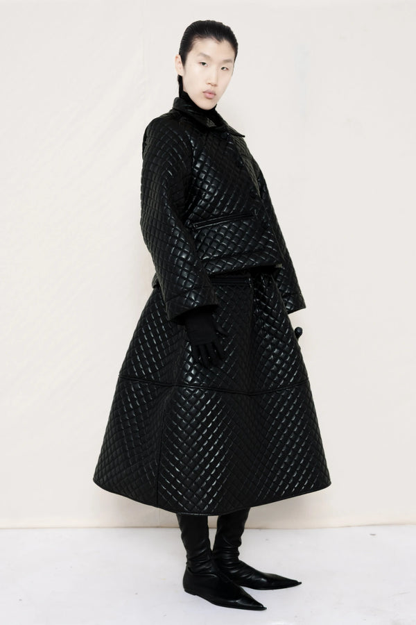 PERSON-WEARING-BLACK-QUILTED-A-LINE-SHIRT-JACKET-WITH-MATCHING-QUILTED-A-LINE-SKIRT-ACCESSORIZED-WITH-BLACK-FLAT-POINTY-BOOT-AND-PIERCED-BLACK-BANANA-TOTE-BAG-THREE-QUARTER-VIEW