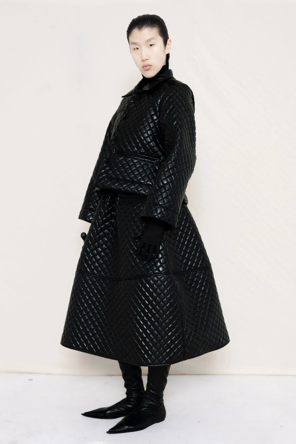woman-wearing-a-black-quilted-jacket-over-a-black-quilted-a-line-calf-length-skirt-and-black-leather-pointed-boots-and-studded-black-leather-banana-bag-on-a-white-background-full-body-three-quarter-view