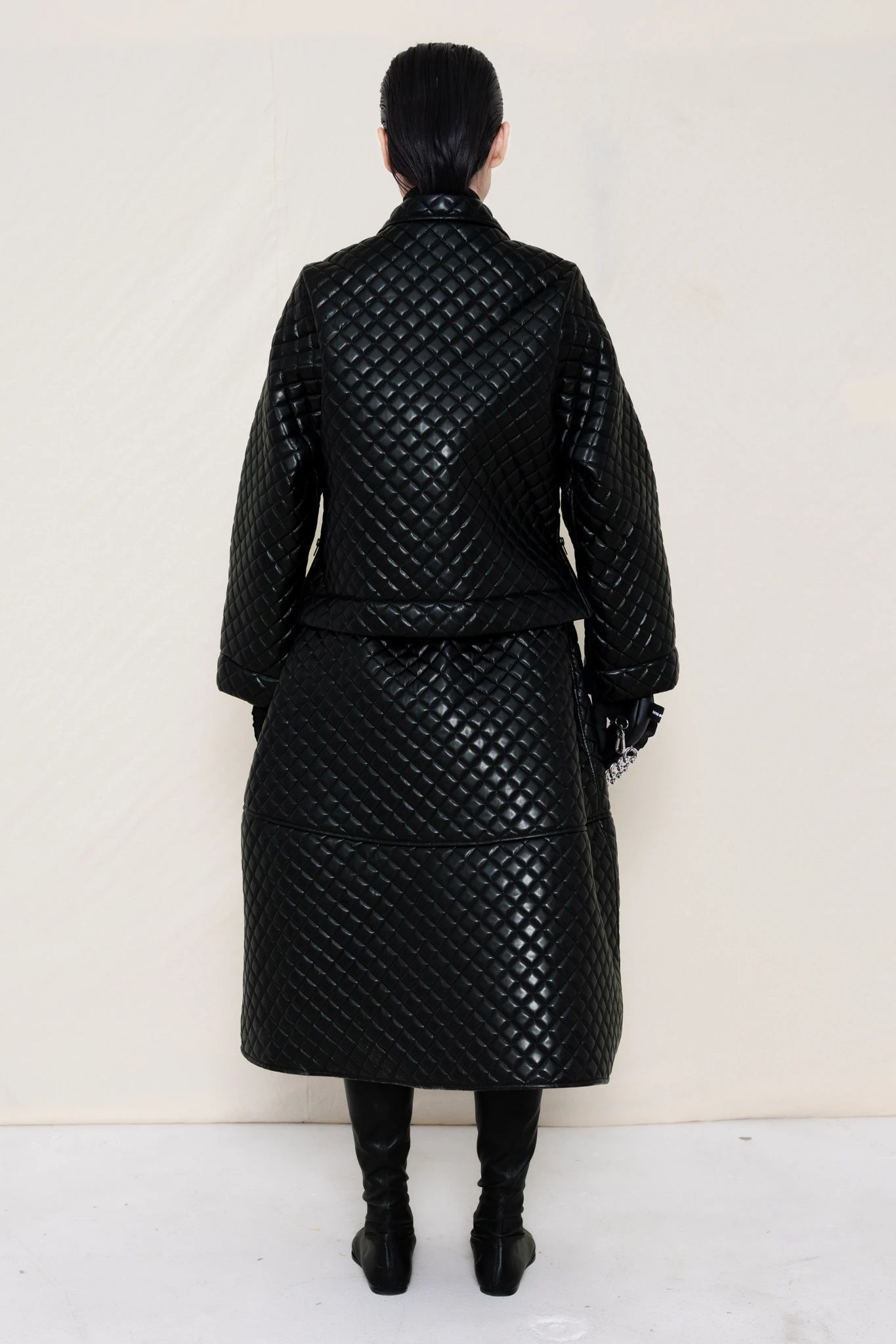 PERSON-WEARING-BLACK-QUILTED-A-LINE-SHIRT-JACKET-WITH-MATCHING-QUILTED-A-LINE-SKIRT-ACCESSORIZED-WITH-BLACK-FLAT-POINTY-BOOT-AND-PIERCED-BLACK-BANANA-TOTE-BAG-BACK-VIEW