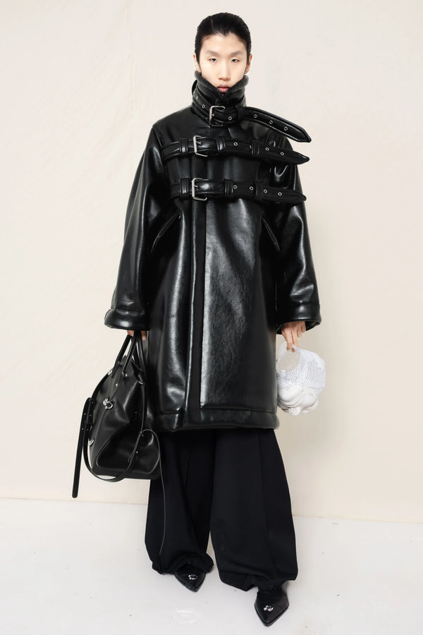 Person-wearing-a-black-shearling-oversized-coat-with- strap-and-buckle-details-worn-over-oversized-black-trousers-paired-with-a-large-black-leather-bag-and-black-pointed-leather-shoes-full-body-front-view