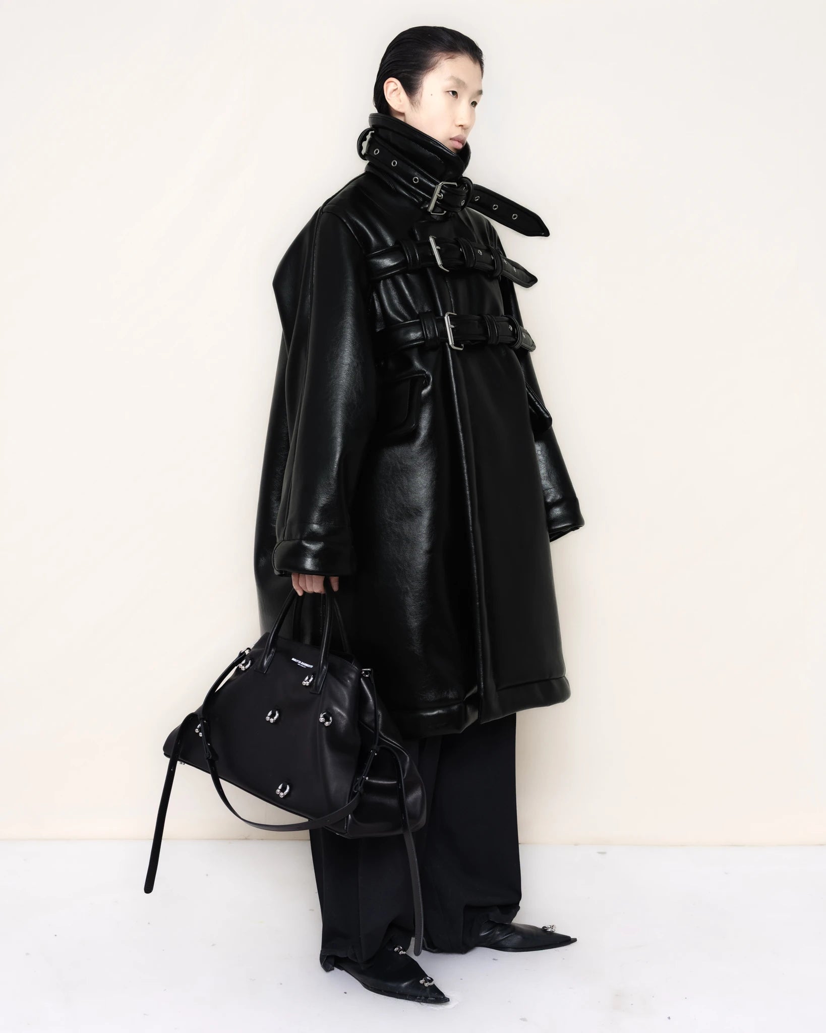 Person-wearing-a-black-shearling-oversized-coat-with- strap-and-buckle-details-worn-over-oversized-black-trousers-paired-with-a-large-black-leather-bag-and-black-pointed-leather-shoes-full-body-three-quarters-view