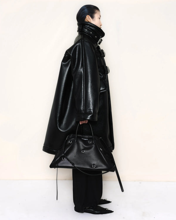 Person-wearing-a-black-shearling-oversized-coat-with- strap-and-buckle-details-worn-over-oversized-black-trousers-paired-with-a-large-black-leather-bag-and-black-pointed-leather-shoes-full-body-side-view