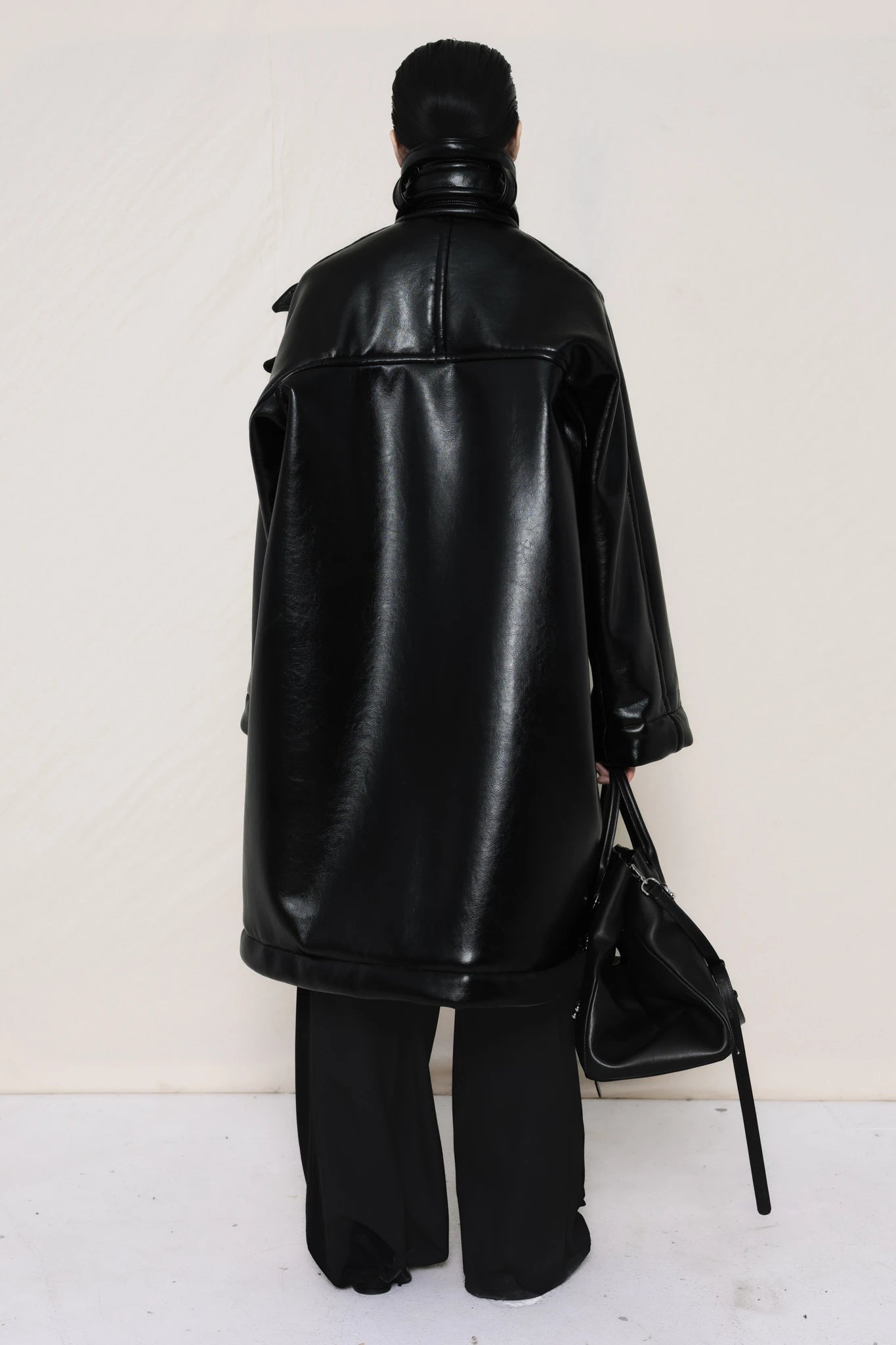 Person-wearing-a-black-shearling-oversized-coat-with- strap-and-buckle-details-worn-over-oversized-black-trousers-paired-with-a-large-black-leather-bag-and-black-pointed-leather-shoes-full-body-back-view