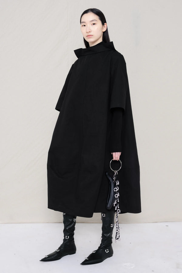 Person wearing a black cape and holding a black handbag against a plain background